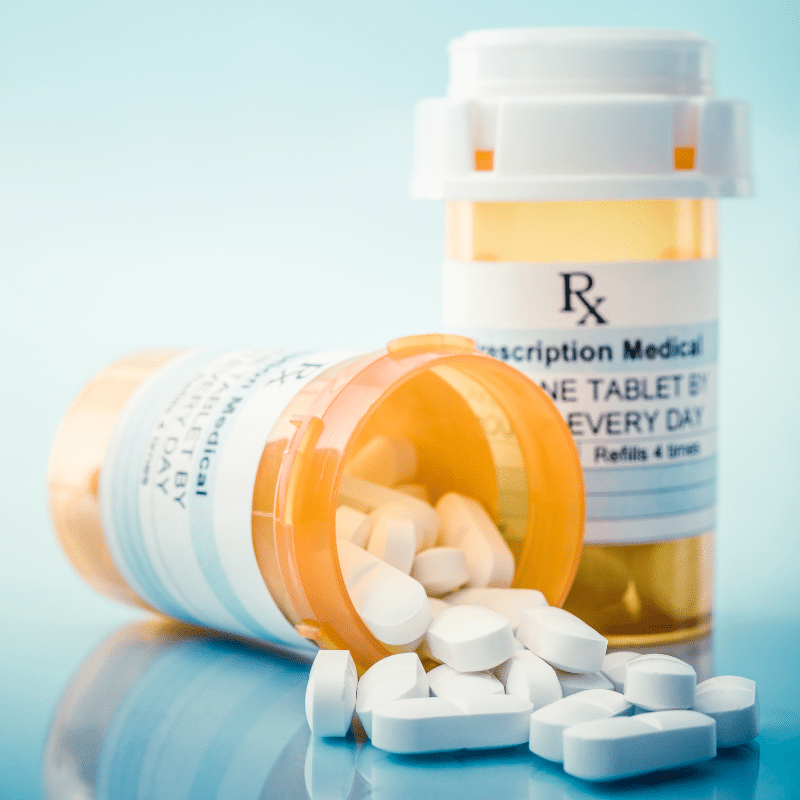 Two prescription pill bottles, one upright and one tipped over with white tablets spilling out onto a smooth surface, against a light blue background.