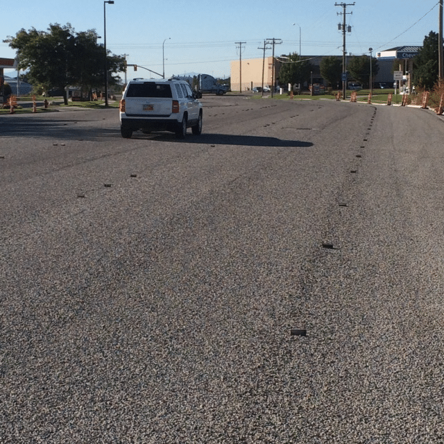 A white SUV drives down an empty, wide urban street on a sunny day. Small, raised pavement markers are visible on the road. A few buildings and trees line the roadside with some construction cones on the right side.