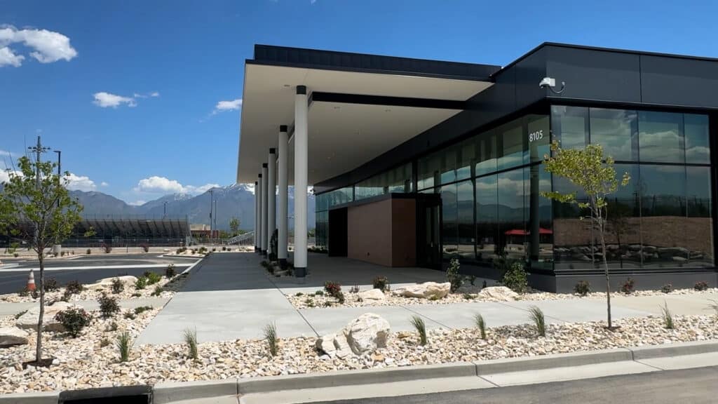 A modern, glass-fronted building with white pillars and a flat roof sits beside a landscaped area with rocks and small plants under a blue sky with mountains in the background.