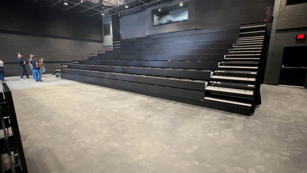 A wide view of an empty, modern black box theater with black tiered seating, a concrete floor, and a few people standing on the left. A windowed control booth overlooks the space from the back wall.