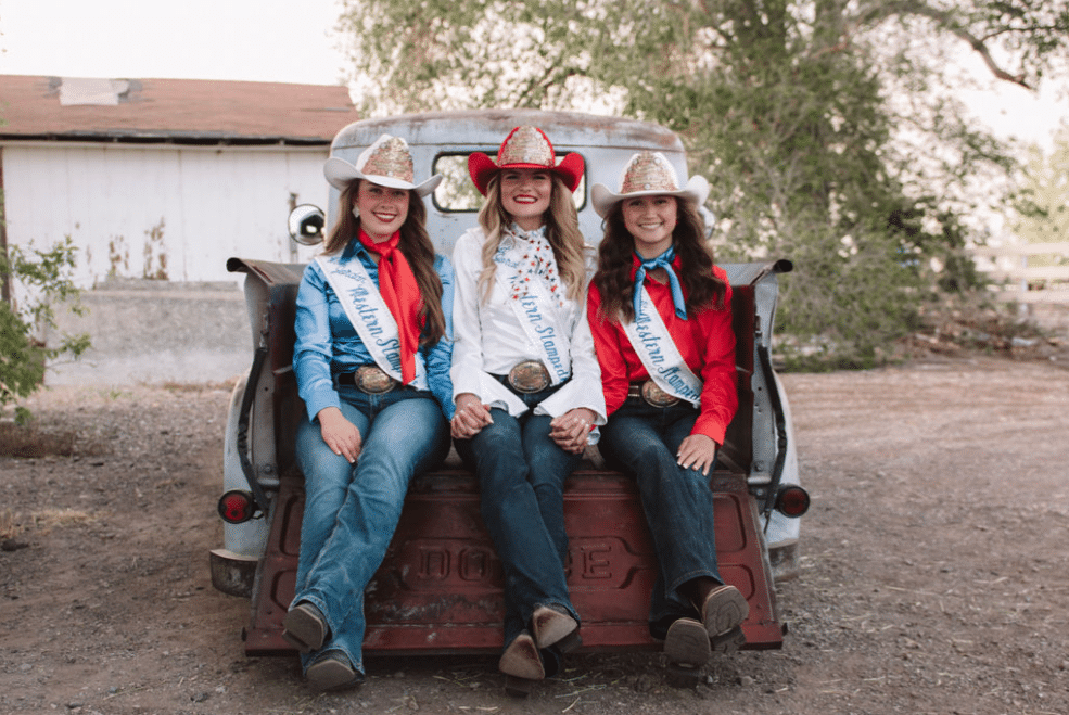 Three women in western outfits, sashes, and cowboy hats sit on the open tailgate of a vintage Dodge truck outdoors, smiling and holding hands with a barn and trees in the background.