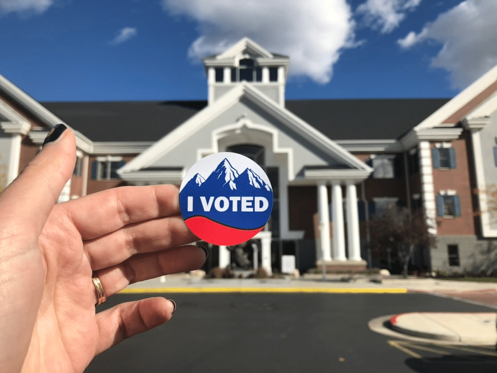 A hand holds a circular I VOTED sticker with a mountain design in front of a large building with columns and a peaked roof under a partly cloudy sky.