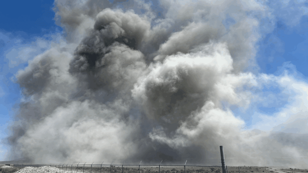 A massive cloud of dark gray smoke billows into the sky above a fenced, barren landscape under a bright blue sky, suggesting a large fire or explosion in an open area.