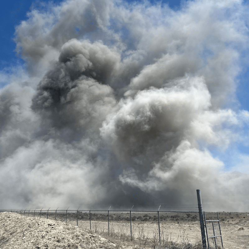 A large, dense cloud of dark gray smoke billows into the sky behind a barbed wire fence in a dry, barren landscape under a blue sky.