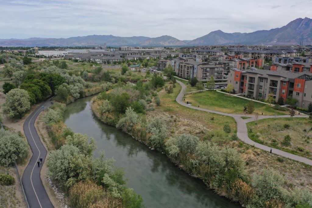 A winding river flows through a green park with walking paths, surrounded by trees and modern apartment buildings; mountains and a cloudy sky appear in the background.