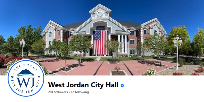 A large American flag hangs on the front of a red and white building with columns, identified as West Jordan City Hall. Trees, lamp posts, and brick walkways are in the foreground under a clear blue sky.