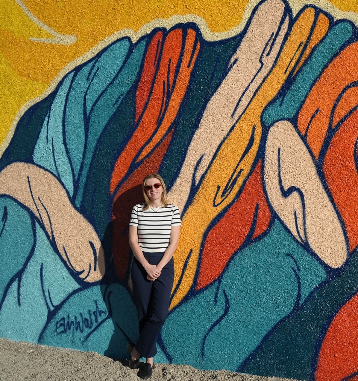 A woman wearing sunglasses, a striped shirt, and dark pants stands and smiles in front of a colorful mural featuring abstract shapes and bold, flowing lines in blue, orange, and yellow tones.