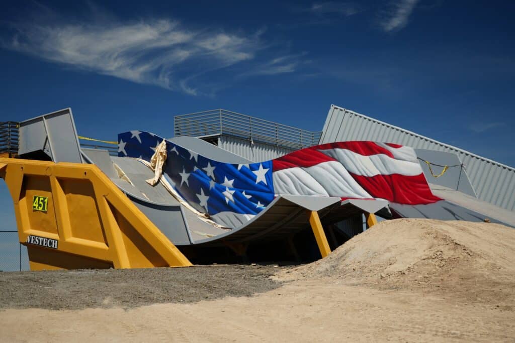 A large, crumpled American flag installation lies on top of a yellow industrial container with sand piles nearby, set against a blue sky.