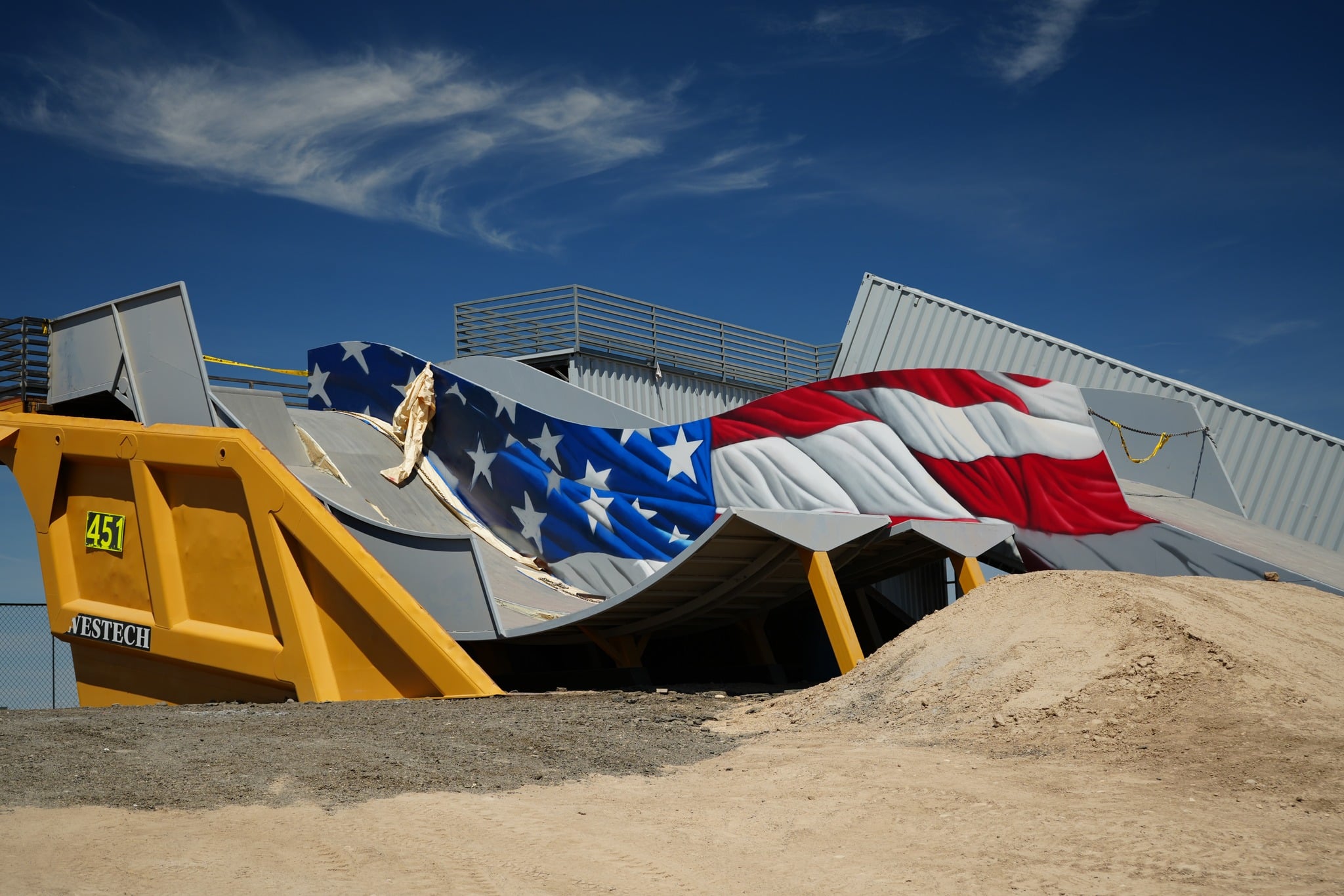 A large, crumpled American flag installation lies on top of a yellow industrial container with sand piles nearby, set against a blue sky.