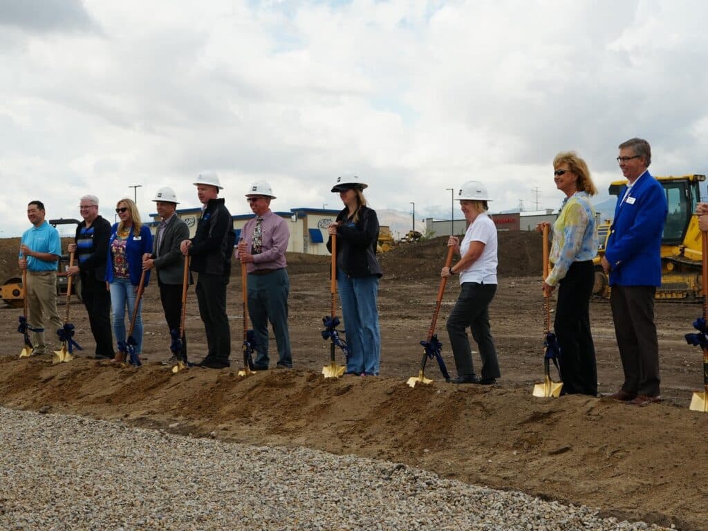 A group of people, some wearing hard hats, stand in a row at a construction site, each holding a shovel planted in dirt, participating in a groundbreaking ceremony with construction equipment in the background.
