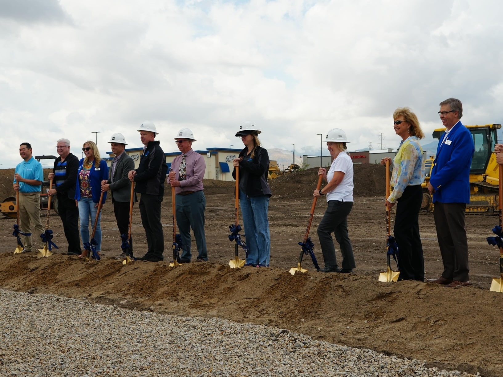 A group of people, some wearing hard hats, stand in a row at a construction site, each holding a shovel planted in dirt, participating in a groundbreaking ceremony with construction equipment in the background.