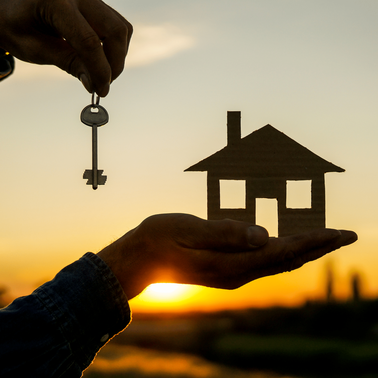 A person holds a cardboard cutout of a house in one hand and a key in the other against a sunset sky, symbolizing home ownership or real estate.