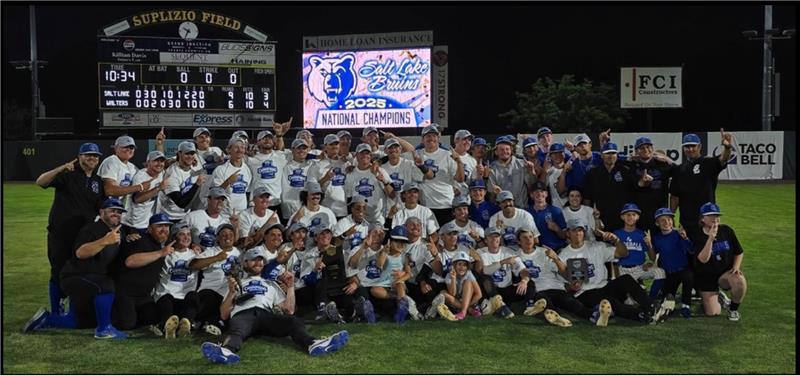 A large softball team wearing white National Champions shirts and hats poses on a baseball field at night, smiling and holding up number-one gestures in front of a scoreboard showing their victory.