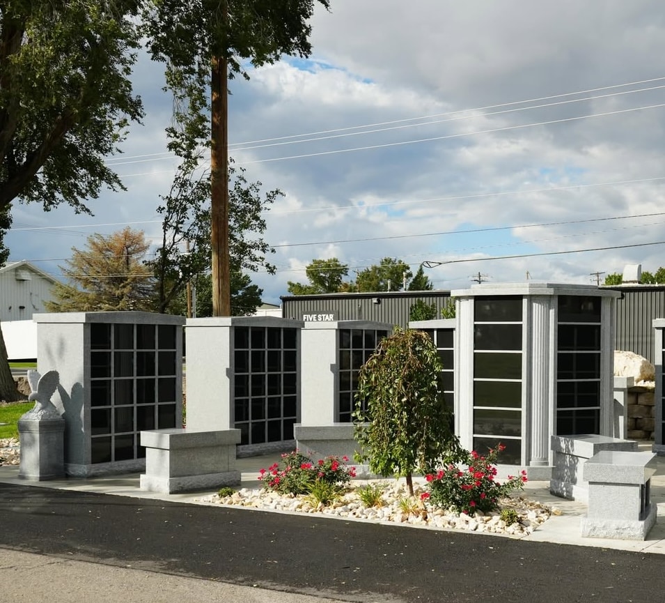 A cemetery columbarium with several gray granite structures featuring glass-front niches, surrounded by benches, a small flowering garden, and trees under a partly cloudy sky.