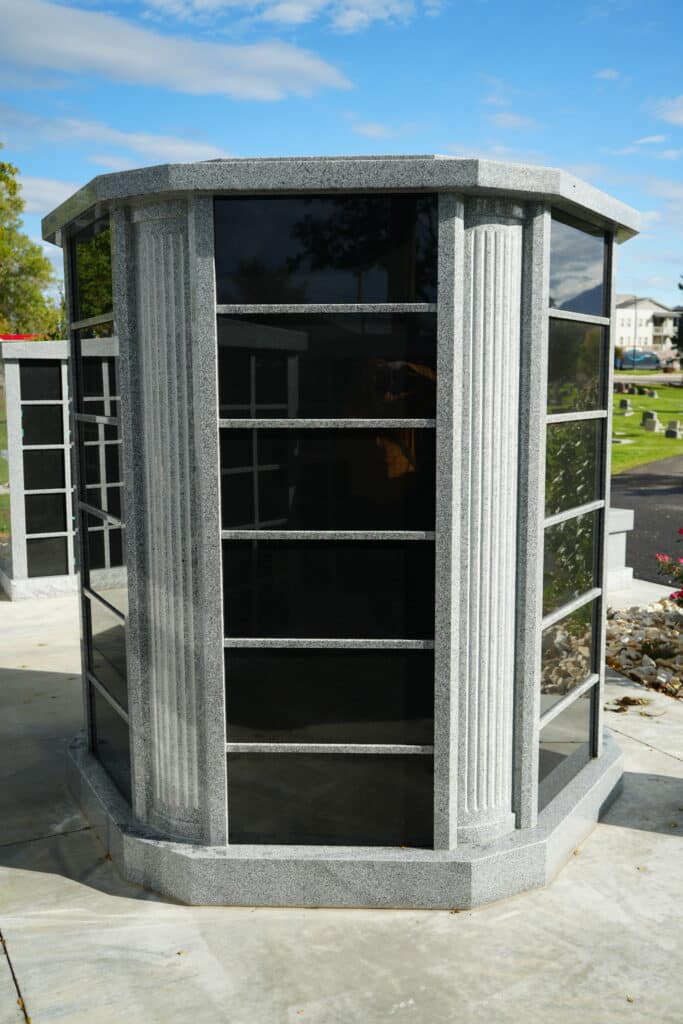 A gray granite columbarium with black glass niches, standing outdoors in a cemetery, surrounded by grass and gravestones under a blue sky.