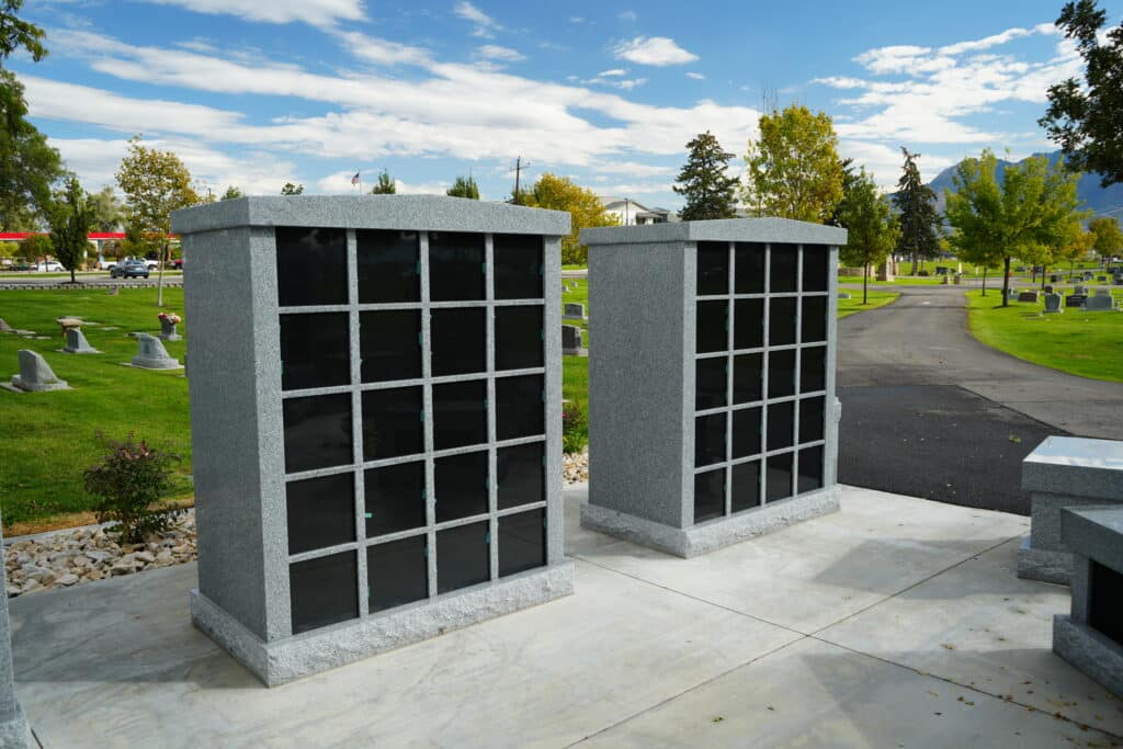 Two granite columbarium structures with black niches stand in a cemetery surrounded by green grass, trees, and gravestones under a partly cloudy sky.