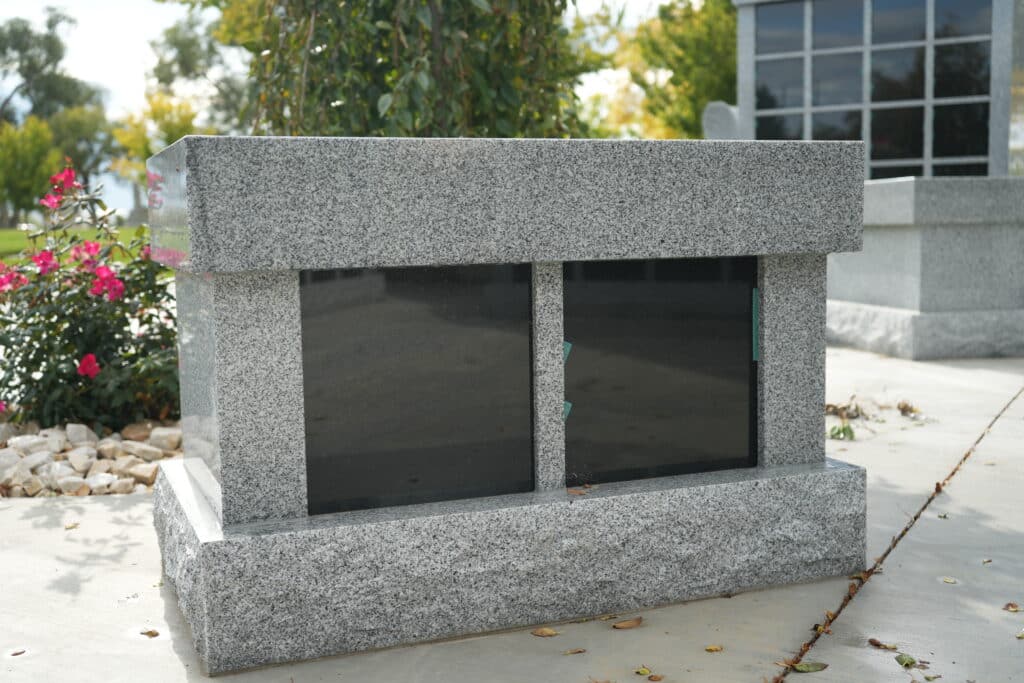 A gray granite double-headstone with two blank black panels stands outdoors, surrounded by some greenery, pink flowers, and white stones in a cemetery setting.