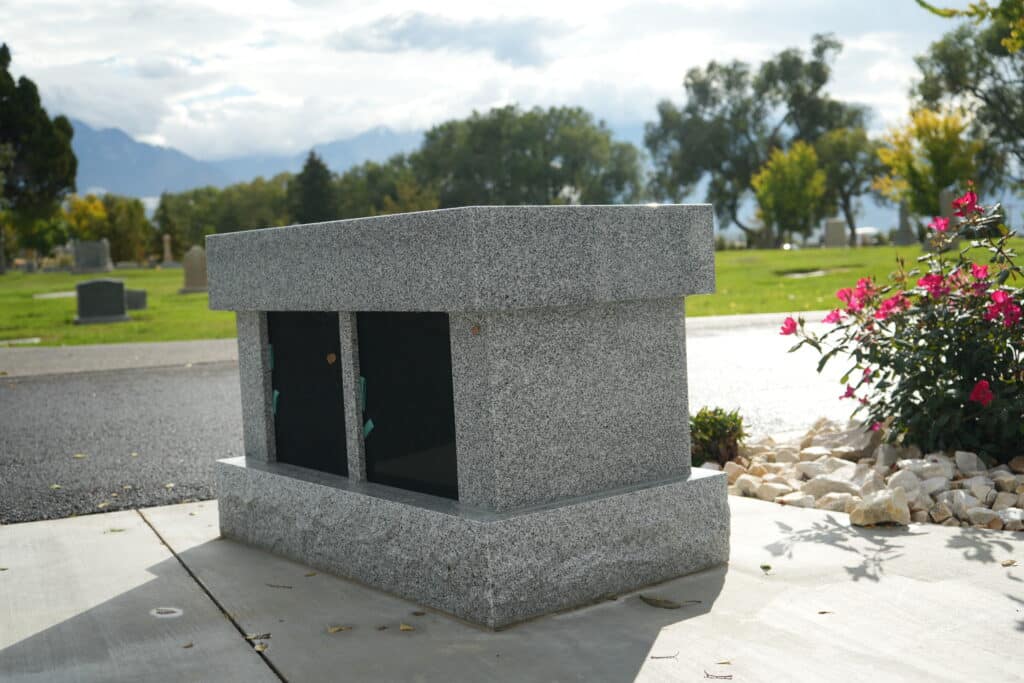 A gray granite columbarium with two niches sits on a concrete slab in a cemetery, surrounded by green grass, trees, and pink flowers under a partly cloudy sky.