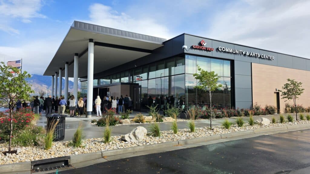 A group of people gathers outside a modern building labeled Canyon View Community & Arts Center, surrounded by landscaped plants, with mountains and flags visible in the background under a partly cloudy sky.