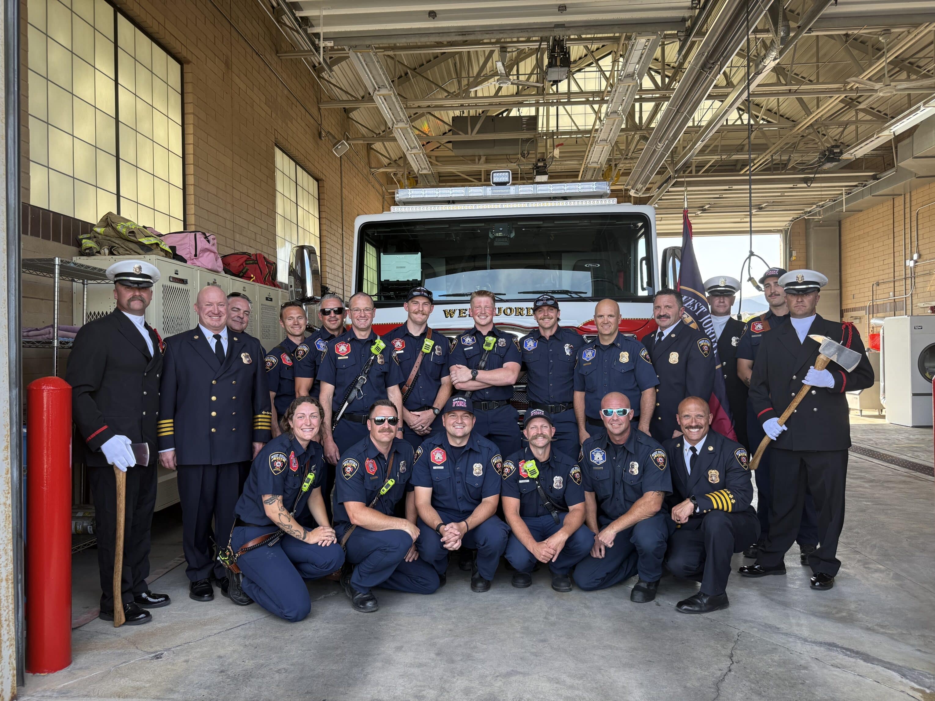 A group of firefighters in uniform pose and smile in front of a fire truck inside a fire station, with firefighting equipment visible around them.