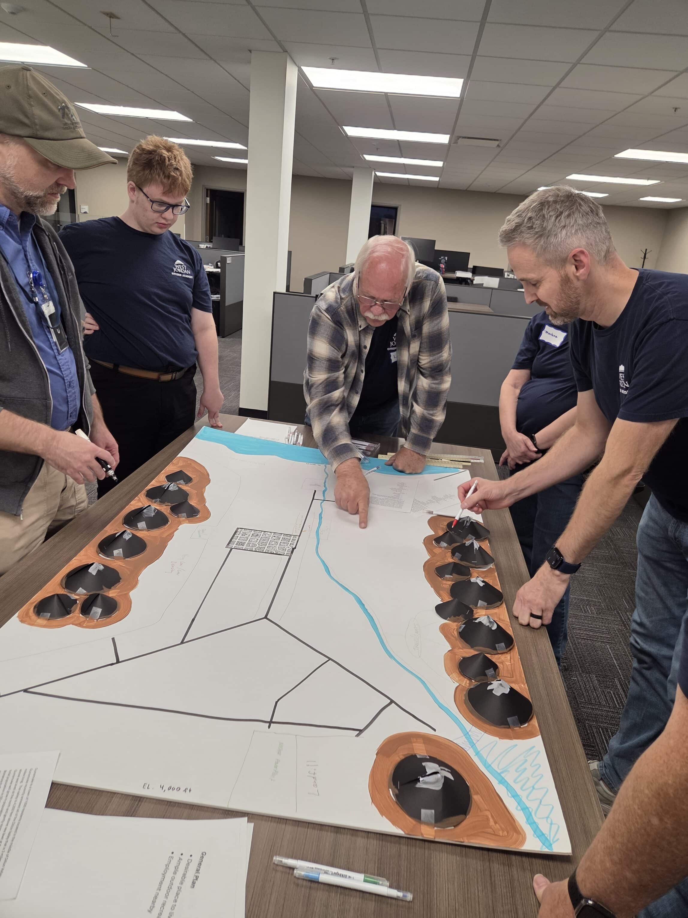 Five people stand around a table, discussing and pointing at a large paper map with models, likely involved in a collaborative planning or design activity in an office setting.