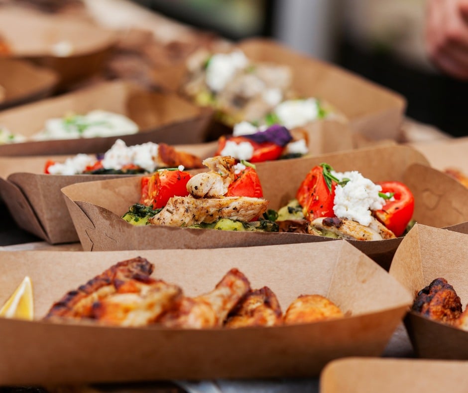 A close-up of several brown paper food trays filled with grilled chicken, roasted vegetables, and dollops of white cheese, arranged on a table. The background is slightly blurred.