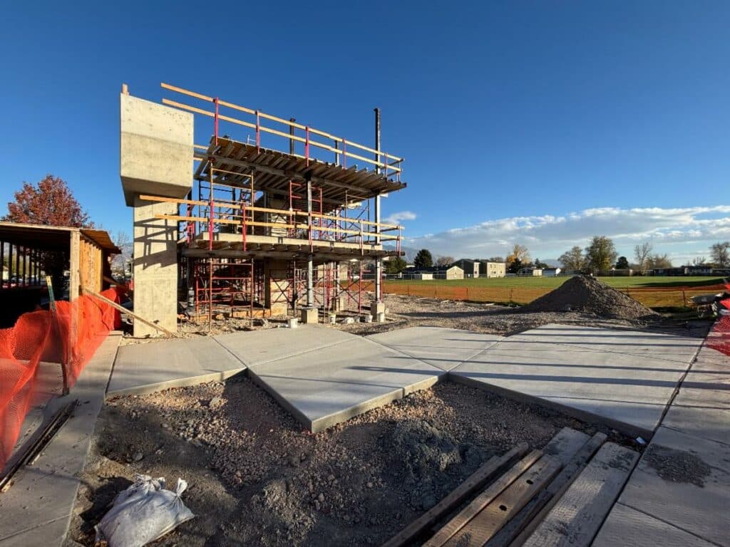 A construction site with a partially built, two-story concrete structure surrounded by scaffolding, fresh pavement, piles of gravel, orange safety fencing, and an open field in the background under a clear blue sky.