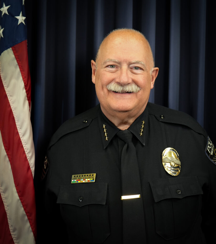 A smiling older man in a black police chief uniform stands next to an American flag, in front of a dark blue curtain background.