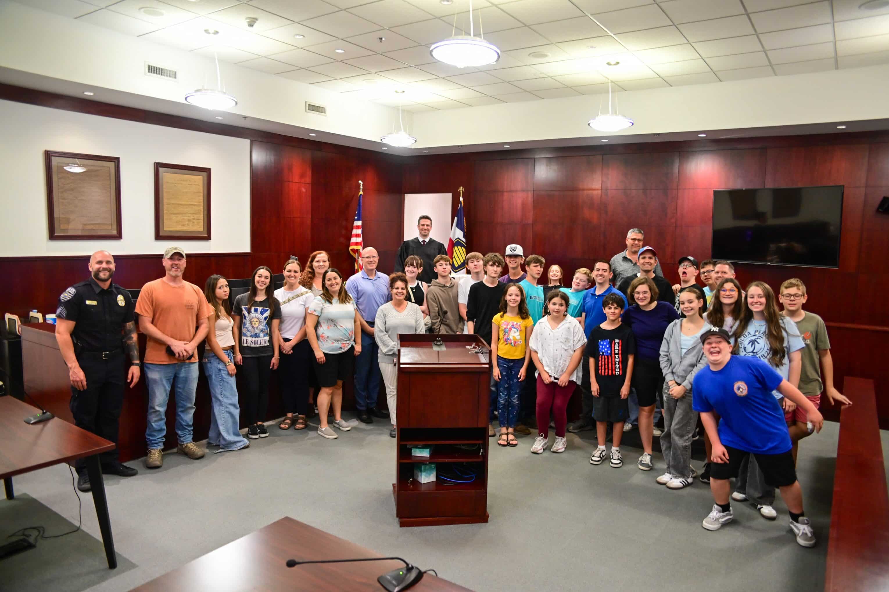 A large group of adults and children pose and smile for a group photo inside a courtroom, standing in front of a judge’s bench, American flags, and wood-paneled walls.