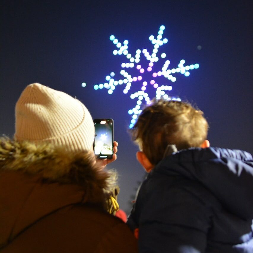 A person in a winter coat and hat holds up a phone to photograph a child in a blue jacket. They both watch a snowflake-shaped drone light display in the night sky.