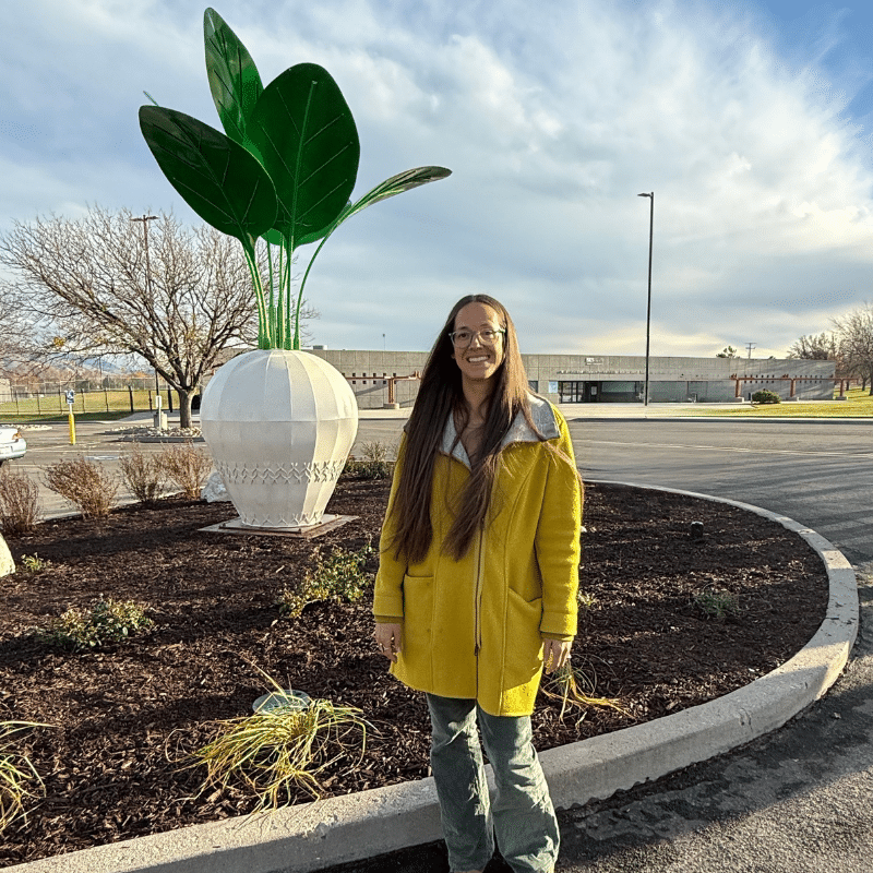 A woman with long brown hair, wearing glasses and a yellow coat, stands smiling in front of a large white radish sculpture with green leaves in a landscaped roundabout on a partly cloudy day.
