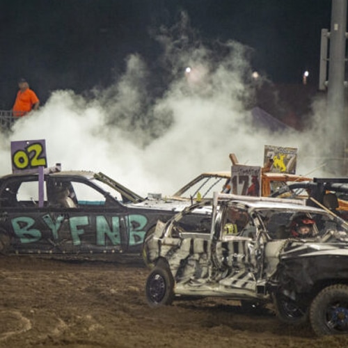 Cars with spray-painted numbers and letters are crashing at a demolition derby, with smoke rising in the background. A person in an orange shirt stands near the arena’s edge, watching the action.