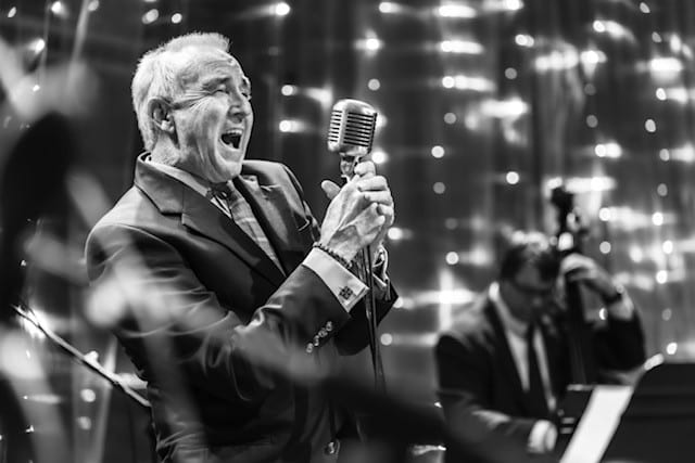 An older man in a suit passionately sings into a vintage microphone on stage, while a musician in the background plays a double bass. The scene is in black and white with sparkling lights behind them.