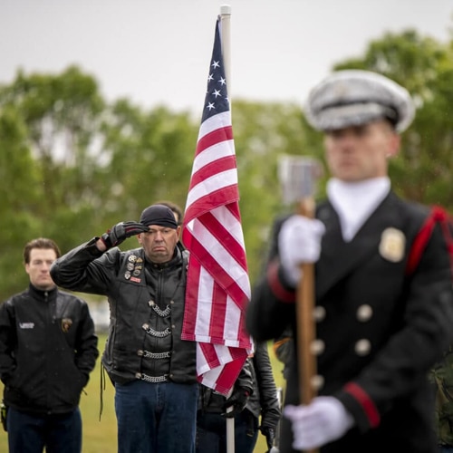 A man in a leather jacket and bandana salutes the U.S. flag outdoors, while other people, including a uniformed person holding a staff, stand nearby. Trees are visible in the background.