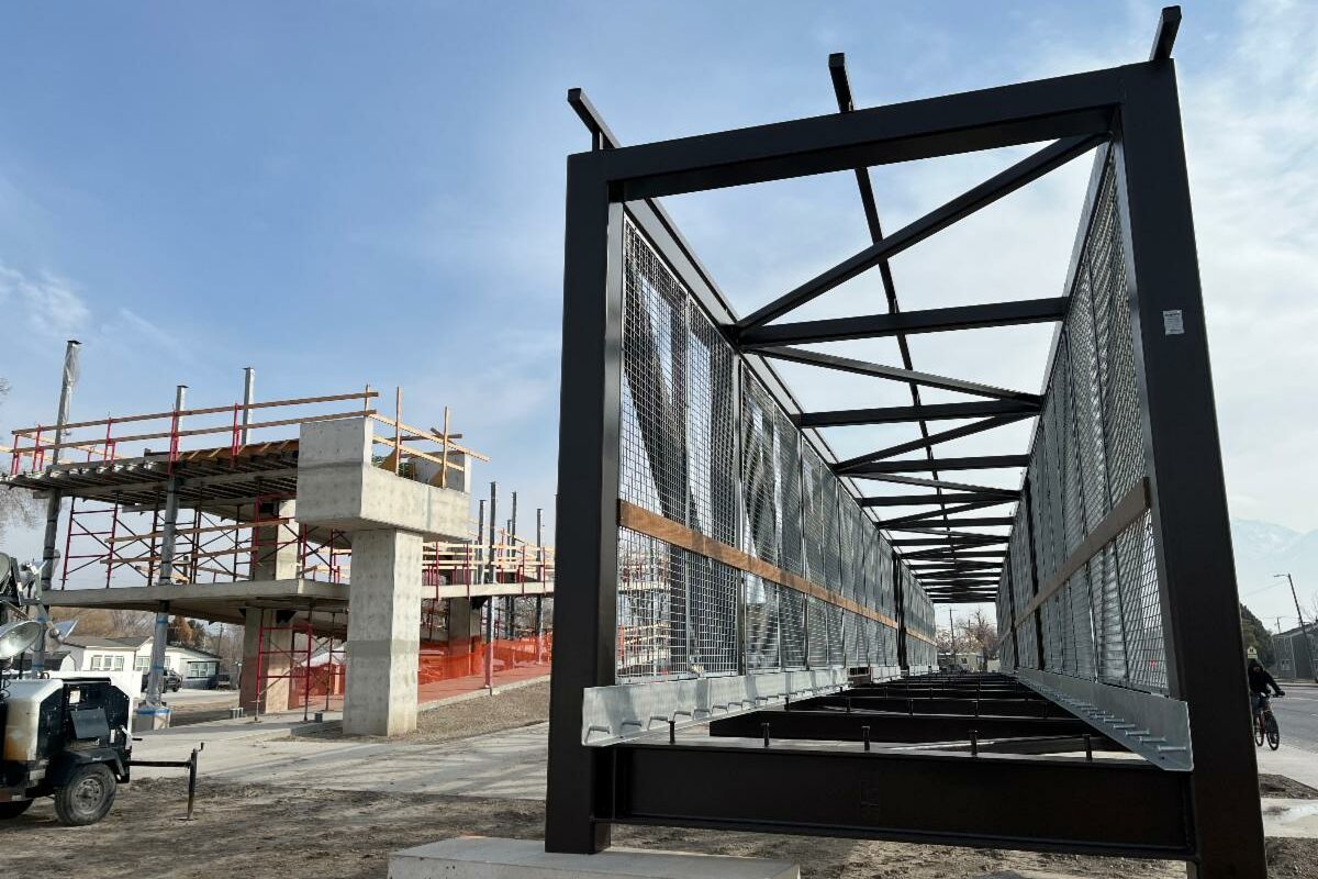 A metal pedestrian bridge under construction stands next to a partially built concrete structure with scaffolding; construction vehicles and equipment are visible on the site under a blue sky.