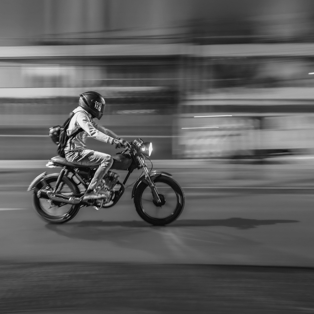 A person wearing a helmet and backpack rides a motorcycle at speed on a city street at night. The background is blurred, emphasizing the motion of the motorcycle. The image is in black and white.