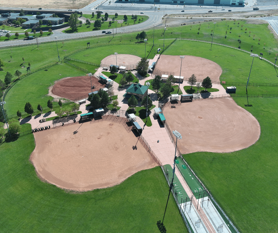 Aerial view of a sports complex with four baseball or softball fields arranged in a cloverleaf pattern, surrounded by green grass, trees, and walking paths, with a parking lot and roads visible in the background.