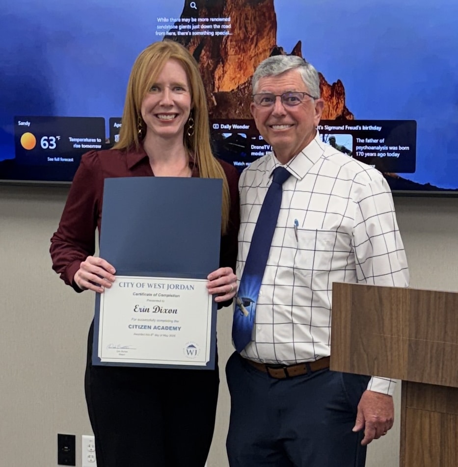 A woman holding a City of West Jordan Citizen Academy certificate stands smiling next to a man in a tie and glasses. They are indoors, with a large screen showing weather and news in the background.