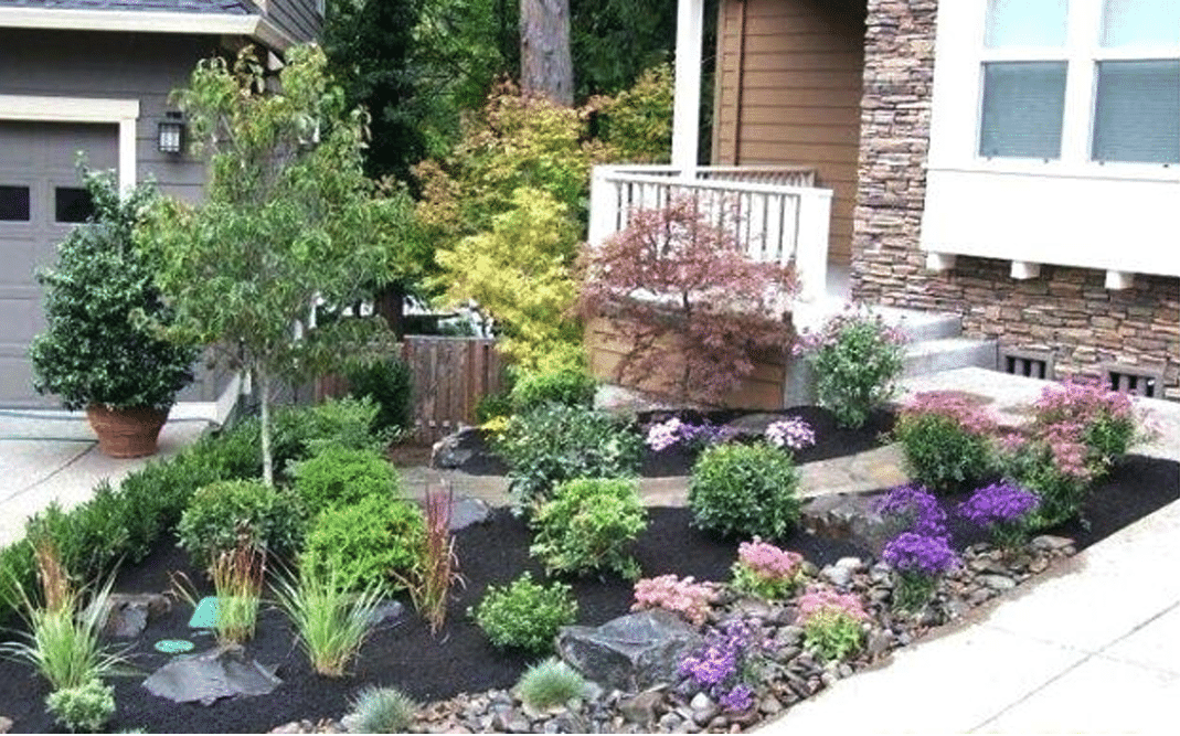 A well-maintained front yard garden with colorful flowers, green shrubs, and small trees bordered by a stone pathway and mulch in front of a house with stone and wood siding.