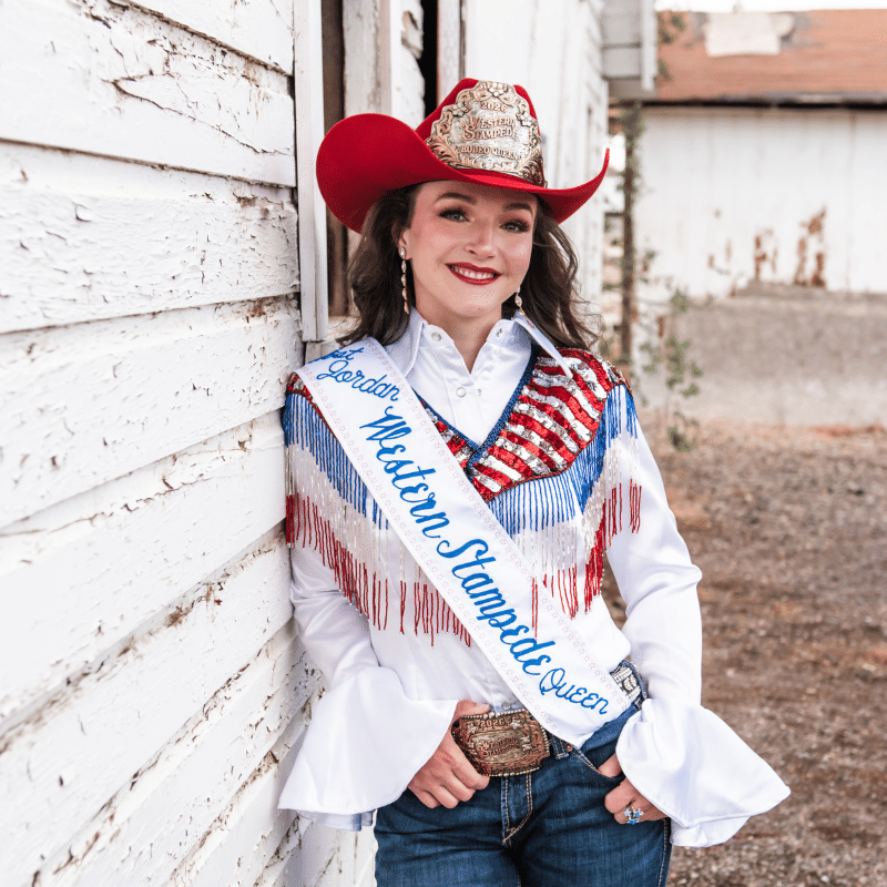 A smiling woman in a red cowboy hat, white shirt with red, blue, and silver fringe, jeans, and a sash reading Western Stampede Queen stands against a worn white wooden wall.