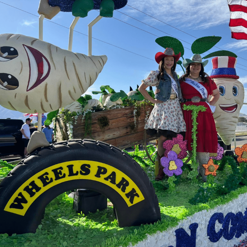 Two women in cowgirl hats and sashes stand on a colorful parade float decorated with vegetables, flowers, and a Wheels Park sign. The float features large cartoon turnips and bright green foliage.
