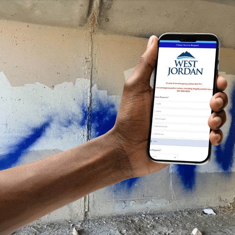 A person holds a phone displaying a West Jordan citizen service report form in front of a concrete wall with blue graffiti.